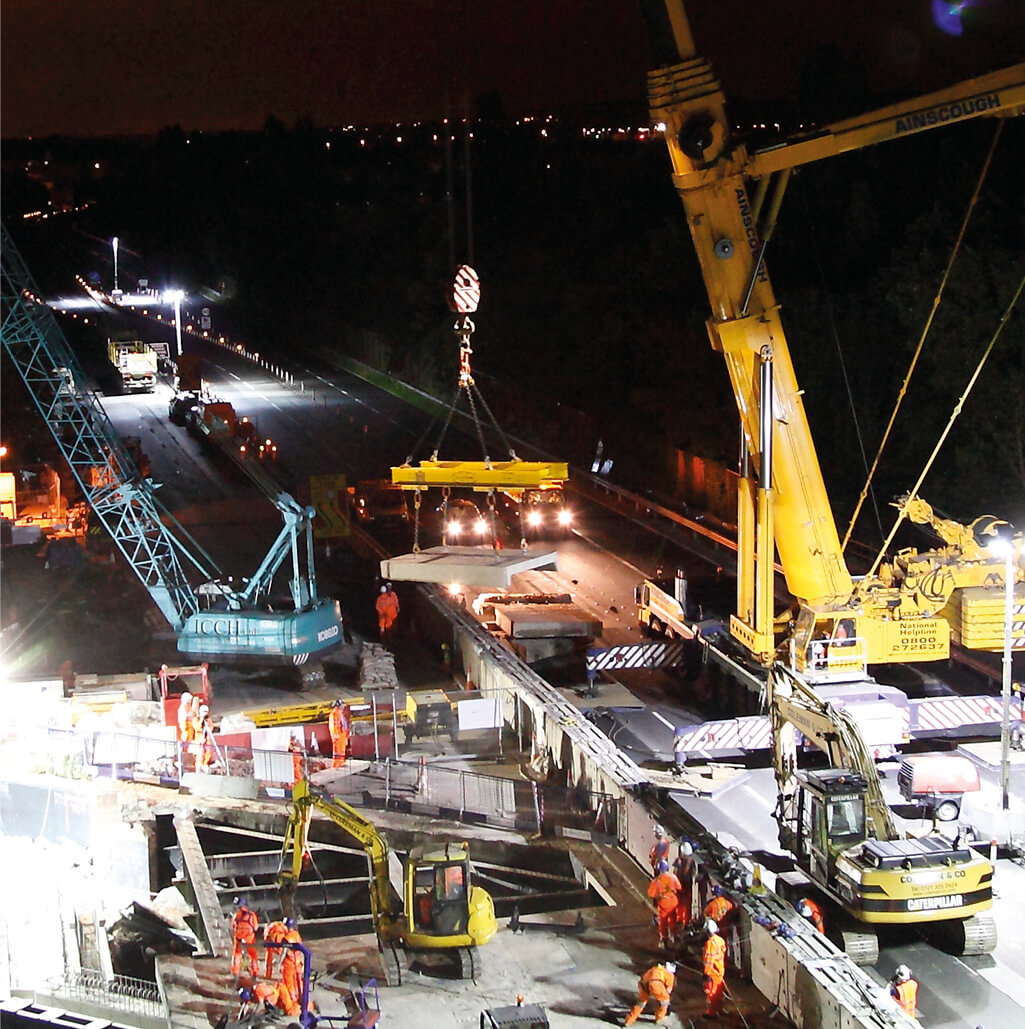 Night scene of bridge removal works. concrete panel is being removed by a Colemans designed lifting panel, which is attached to a mobile crane. Several demolition machinery is in the picture, with working men around the outskirts, wearing protective high visibility wear.