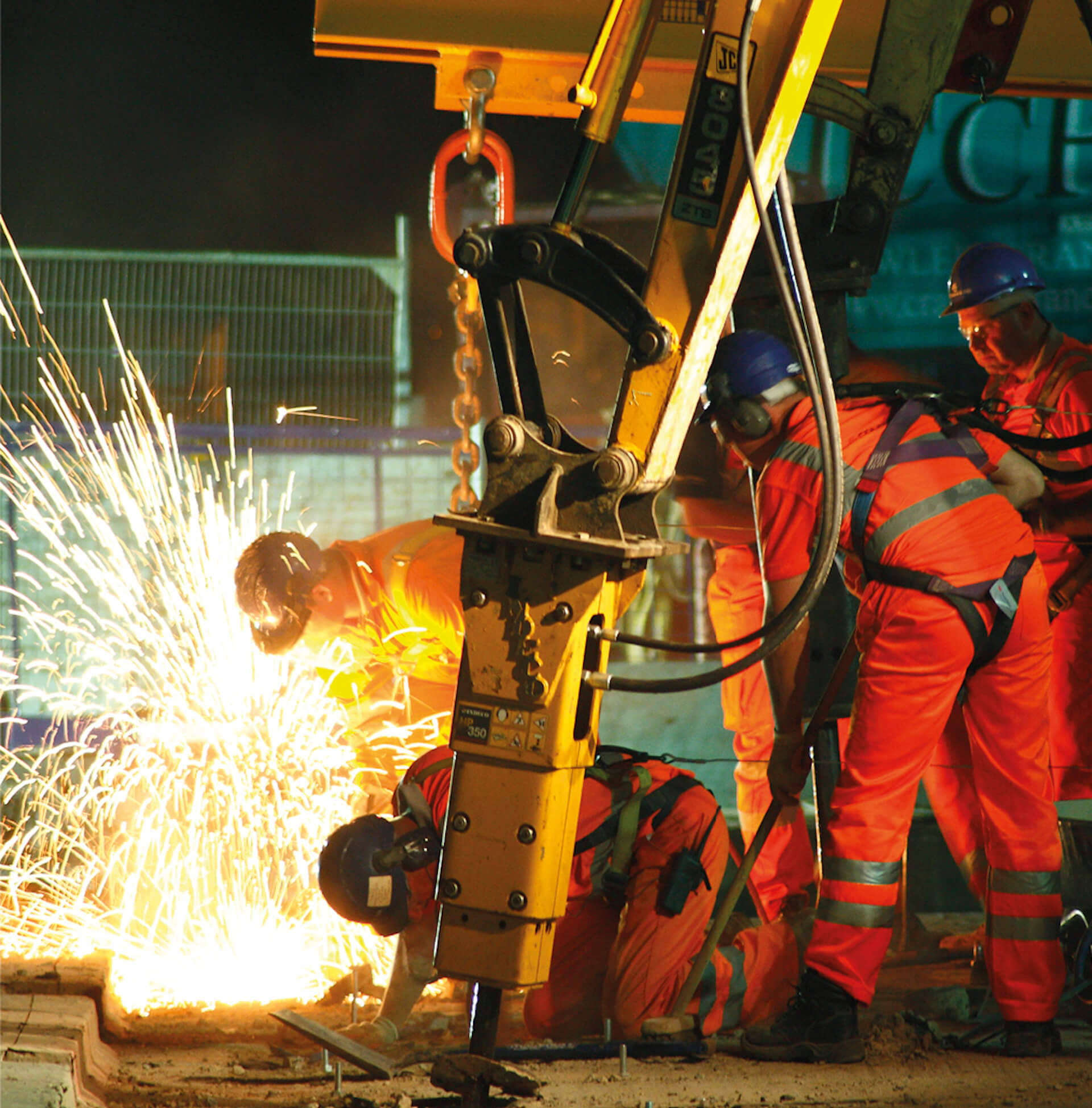 A night scene onsite of concrete being cut by a machine in the foreground. Several workers in the background carrying out hot cutting with sparks flying. Workers wearing protective wear.