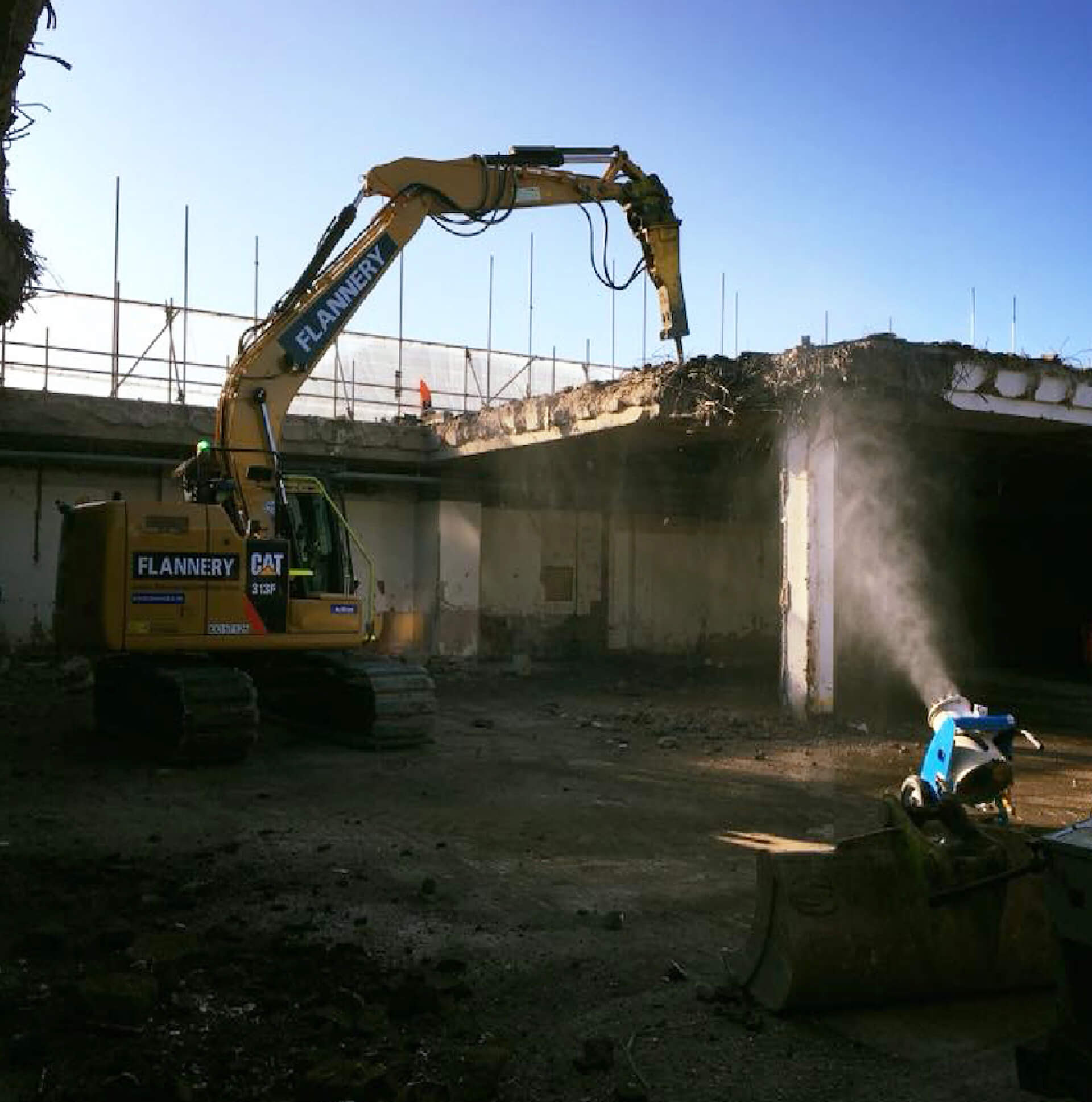 A machine breaking the concrete out of a roof. A water bowser in the foreground to reduce dust.