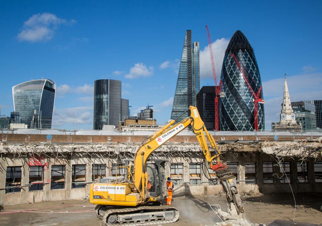 a large piece of rebar and concrete is being munched by a demolition excavator. partially demolished structure is behind the machine. London cityscape is in the background including the Gerkhin and the Shard buildings.