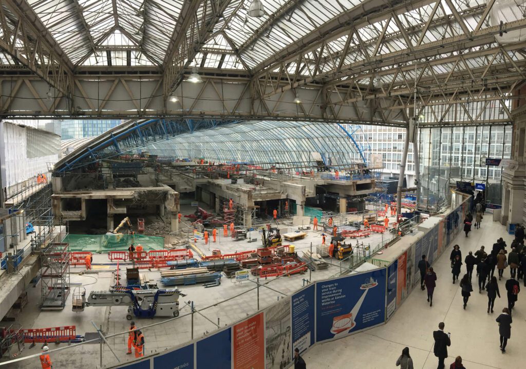 Image of deconstruction site at Waterloo international train station in London. Image shows work in progress on one side of a long hoarding including chapter 8 barriers and workers in safety clothing. The other side of the hoarding shows public walking through the busy train station.