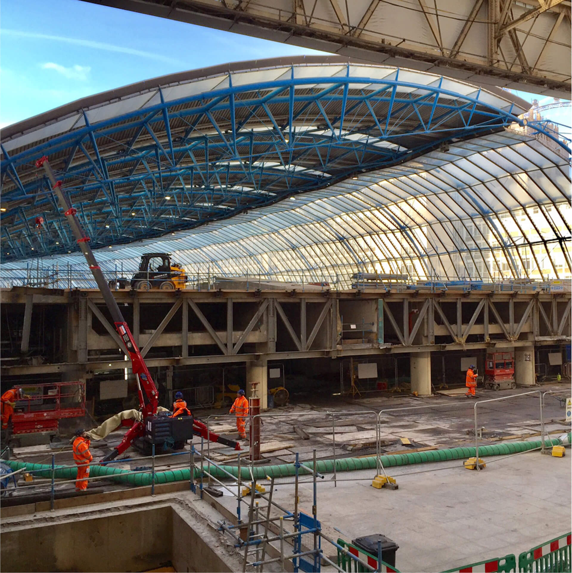 workers removing floor concrete with a machine under overhead roof canopy at Waterloo international train station. Another small machine is positioned on a floor level above in the background.