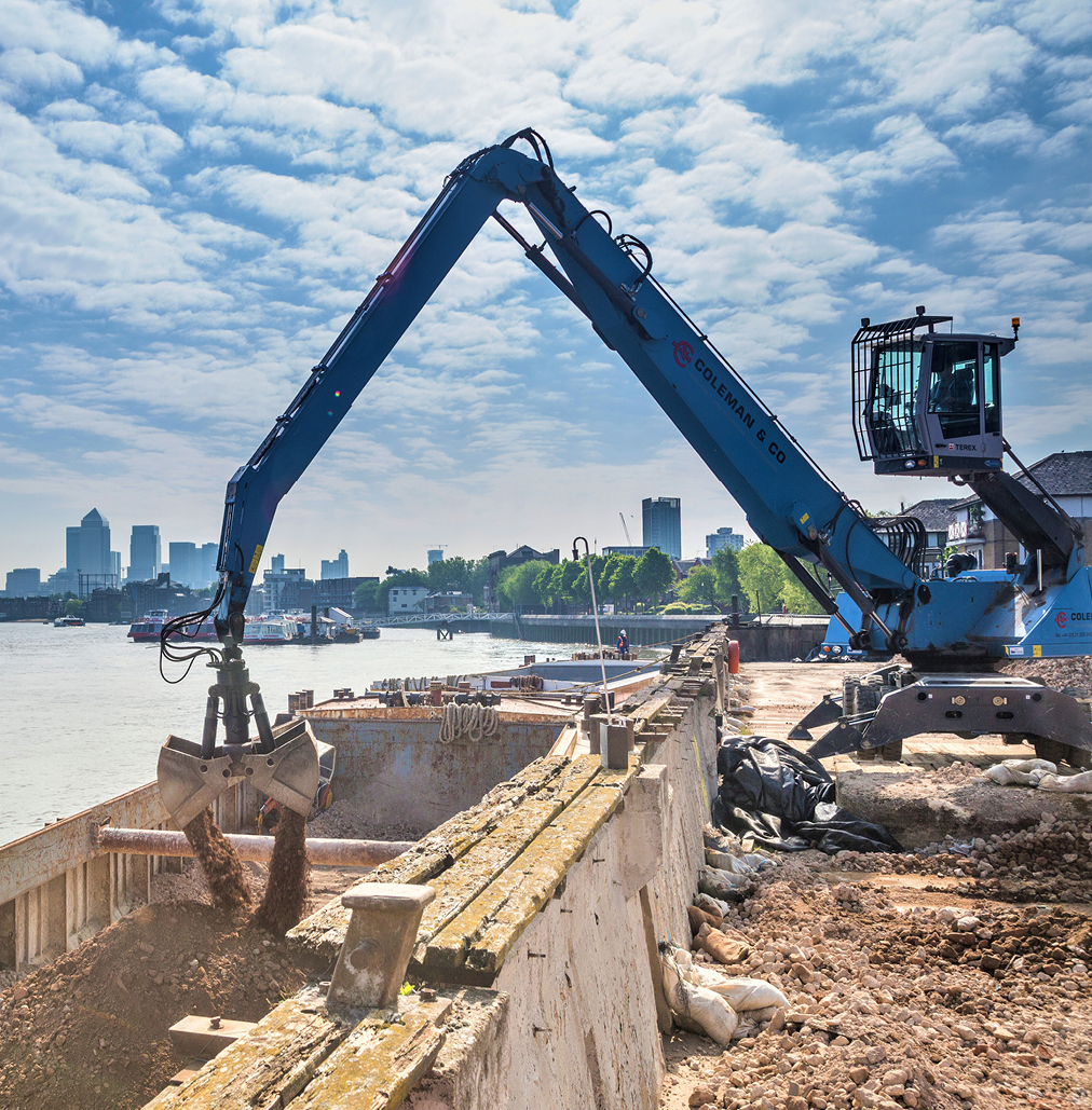 side view of excavator loading contaminated hardcore into a river-boat on the Thames for onward transportation off site. Cityscape and River Thames in the background.