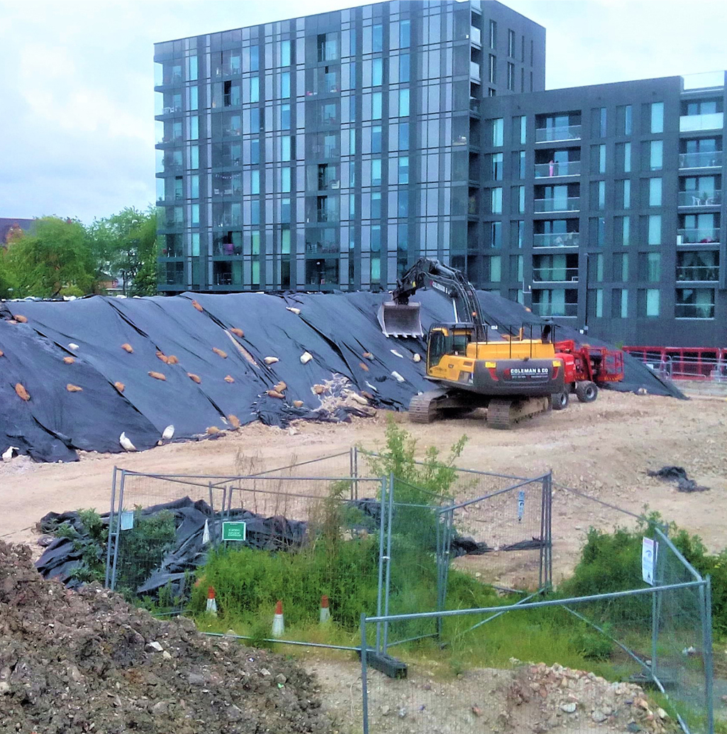 covered stockpiles of ground materials with excavator in the foreground and residential apartment blocks in the background.