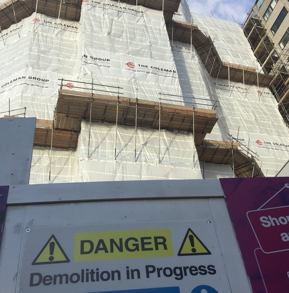 building with scaffolding and protective sheeting with Colemans branding. Hoarding in the foreground with danger - demolition in progress notice board.