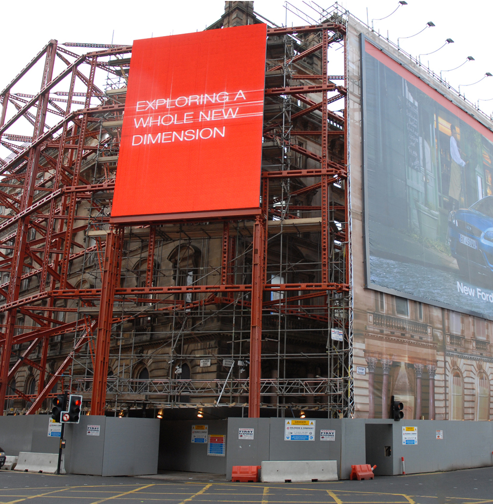 building propped with red metal temporary works structure as part of façade retention. General advertising banner to the front of the building with hoarding all the way around the perimeter.