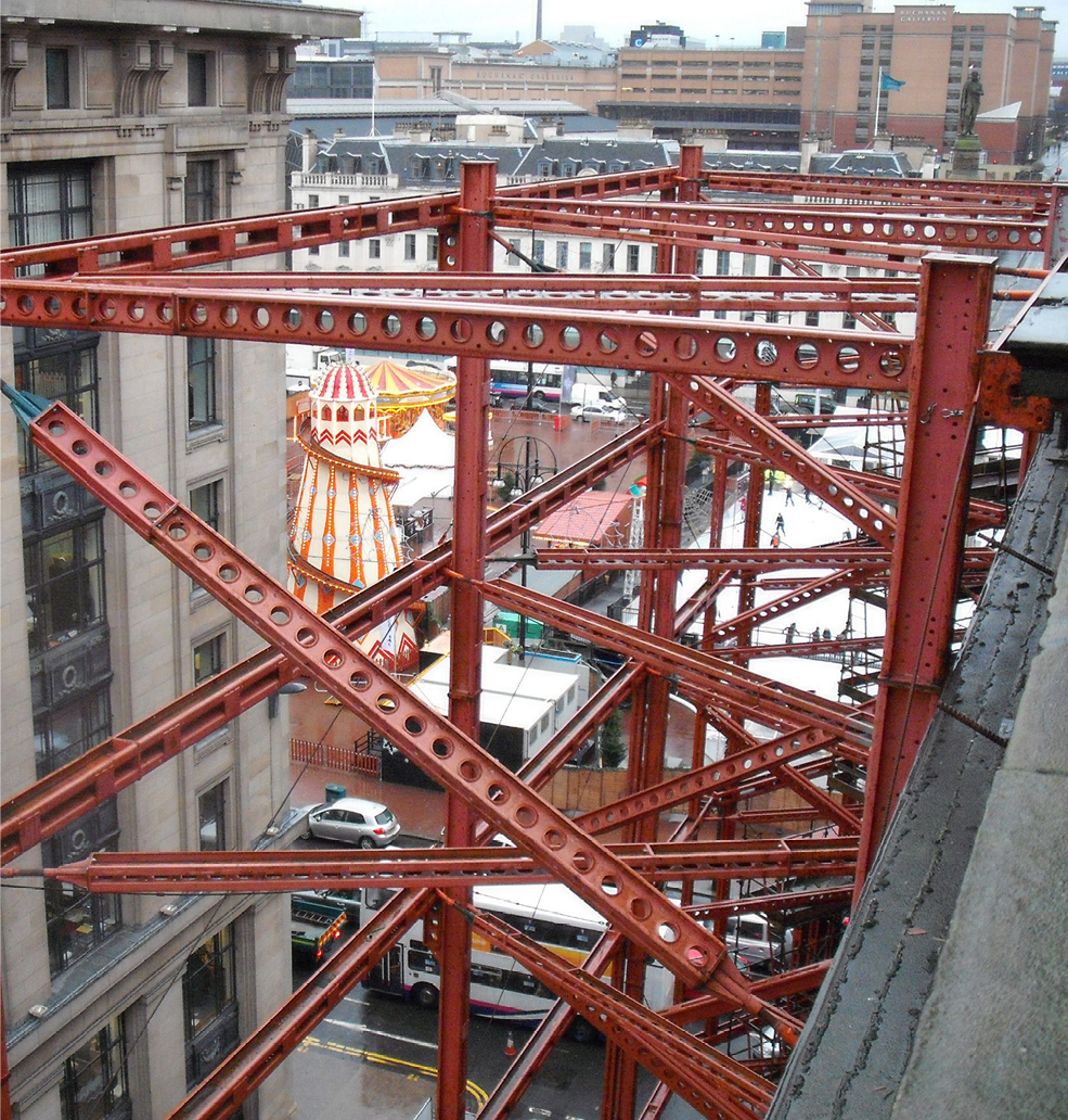 temporary works structure attached to building as part of façade retention works. Red propping at height. Fairground in the background.