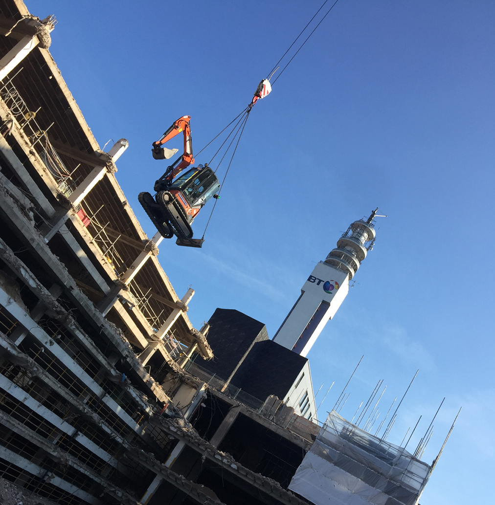 small excavator is being lifted onto site and into position by crane, in front of the partially deconstructed building. Birmingham BT Tower is in the background.