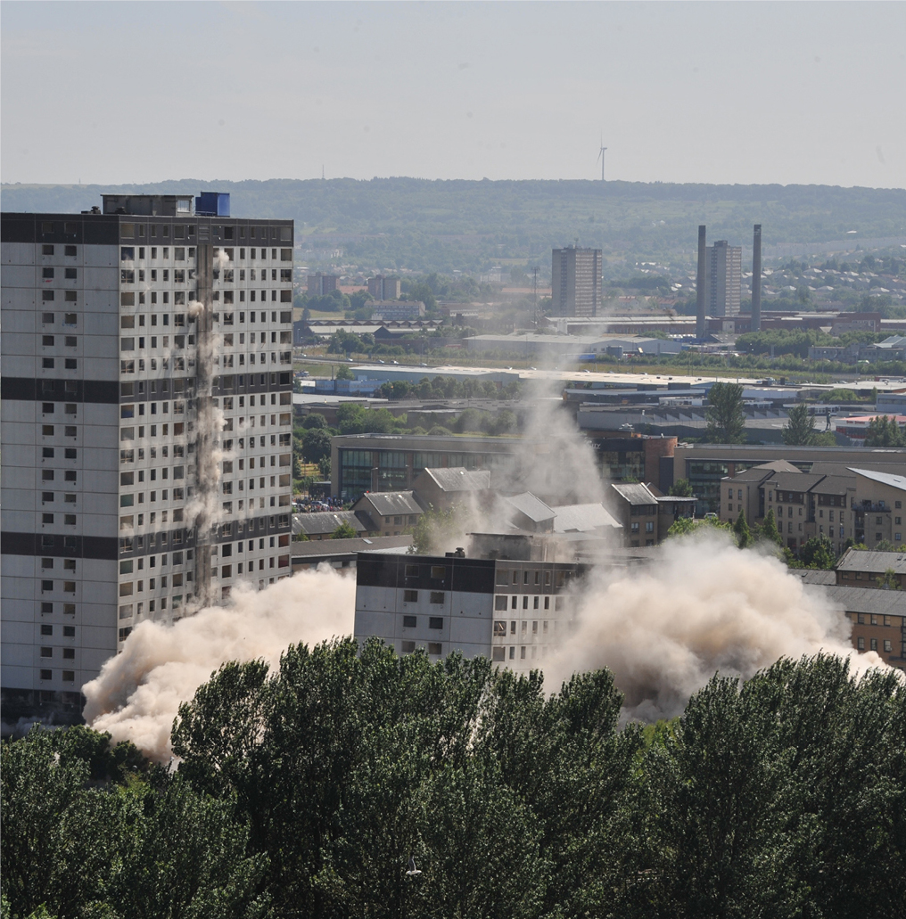Alternative view of the two 24 storey tower blocks coming down during explosive demolition. Dust clouds at either side of the buildings. Trees in the foreground.