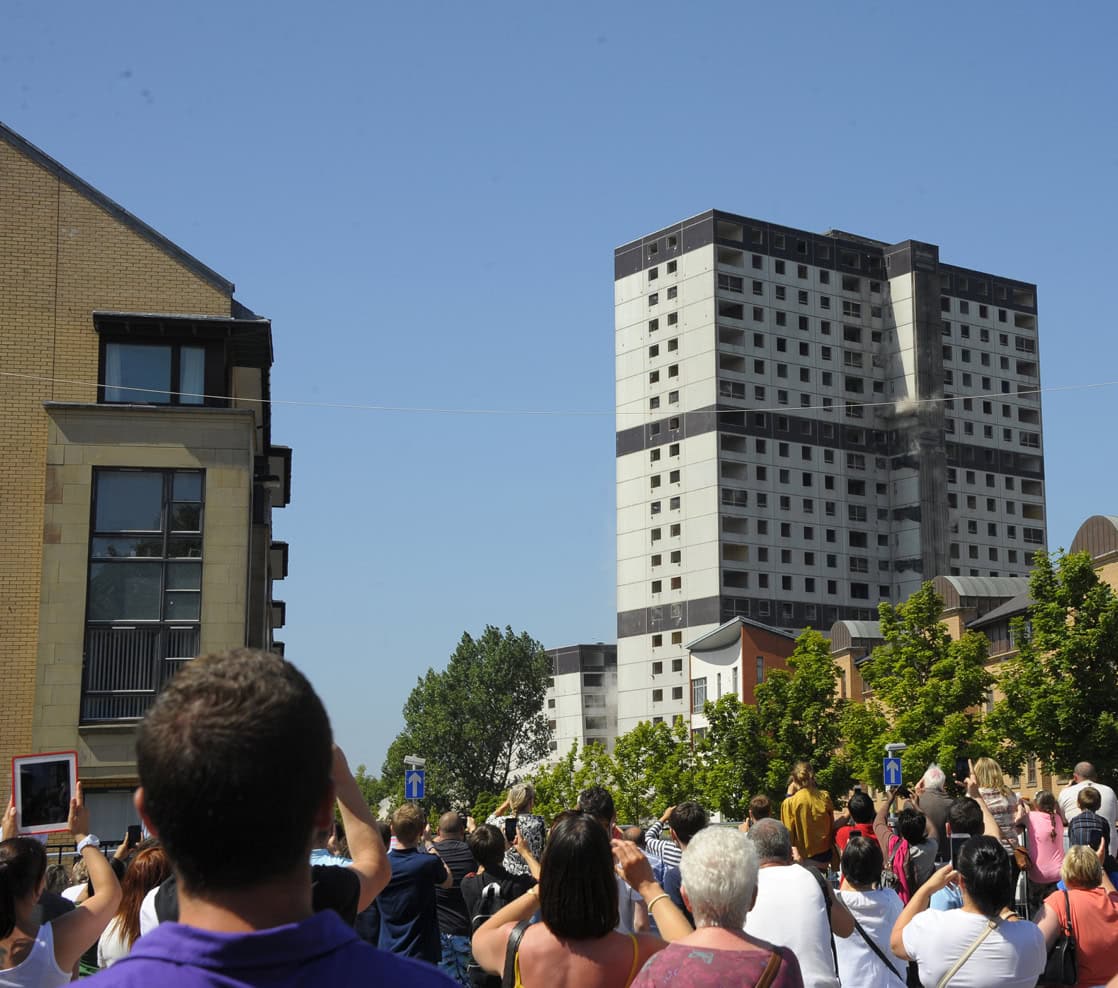 Two 24 storey tower blocks being explosively demolished with a crowd of onlookers in the foreground.